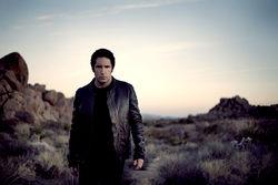 A man dressed in black walking through a desert at sunset.