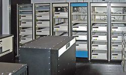  A room with a black box in the foreground and six control cabinet with space for five to six racks each. Most, but not all, of the cabinets are filled with white boxes.