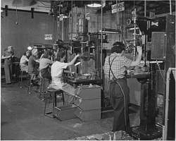 A World War II-era photo of five women working at drill presses, all wearing pants, work shirts, hats, and aprons