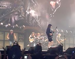 Rock band in performance on a well-lit but hazy stage. At the front is a guitarist; there are two more guitarists, a vocalist off to one side, and a drummer in the rear.