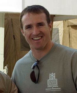 A man is smiling.  He is wearing a gray T-shirt with a pair of black sunglasses hanging from the collar.