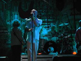 Male singer in white shirtsleeves and trousers, with a band behind him on a small stage.