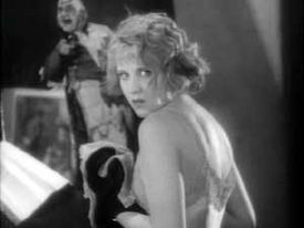 A blond-haired young woman wearing a low-cut garment looks over her left shoulder toward camera. She is wearing thick black gloves and holding an object of unclear nature, possibly made of or covered in cloth. In the background is an illustration of a laughing man, pointing a finger roughly in the woman's direction.