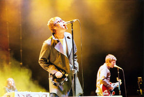 Two-thirds body shot of a singer wearing a coat with wide lapels; a guitar  player is in the background. Both have short, blond hair.