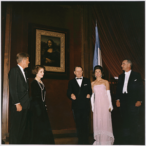 Three men and two women stand near the Mona Lisa. All are dressed formally, one woman in a spectacular pink gown.