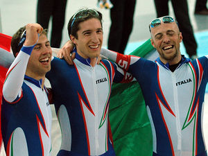 Head and torso photo of three smiling men, wearing glasses on their heads and blue and white jumpsuits with Italia written on the chest. The man in the middle is holding a big Italian flag behind him, while the other two are embracing him.