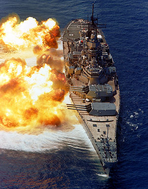 A large warship with guns pointed to the left; fire and smoke can be seen emanating from the gun barrels. Below the ship the dark blue water has taken on a white color owing to the disturbance in wind pressure from the firing of the guns.