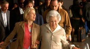 Naomi Watts beaming and facing into soft light holding the arm of an older woman while they take a down escalator at Los Angeles Internation Airport