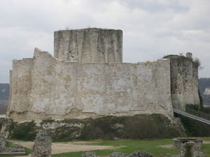 A tall pale grey wall around an enclosure and surrounded by a ditch. A modern metal bridge leads up to the entrance between two towers in the wall. A square stone tower rises above the wall.