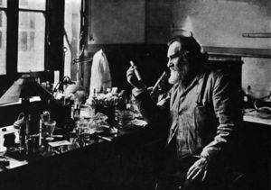 A bearded old man holding up a test tube. He is sitting at a table by a window. The table is covered with many small bottles and test tubes.