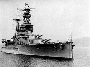 A three-quarter view of a heavily-armoured battleship at anchor. There are two main turrets visible before the bridge, each housing a pair of 15-inch guns. 6-inch guns are housed in a row of individual sideways-facing sponsons. The flank of the ship has a conspicuous bulge at the waterline.