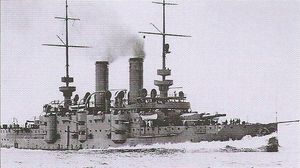 A small grey battleship traveling at full speed with dark smoke billowing out of its two funnels.