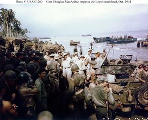 A large crowd of soldiers and jeeps on a beach. There are palm trees in the distance and landing craft offshore. A small group in the center conspicuously wear khaki uniforms and peaked caps instead of jungle green uniforms and helmets.