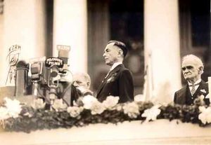 A sepia photo of a dignified debonair man on a decorated balcony who with the many microphones in front of him is about to make an address. Large columns of a building are in the background and a wizened official stands at a distance behind.