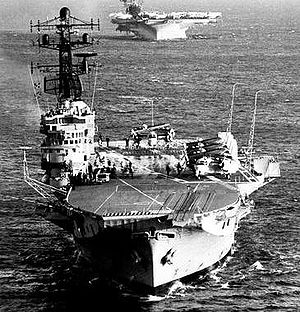 An aerial photo of an aircraft carrier with several aircraft on her flight deck. Another carrier is visible in the background.