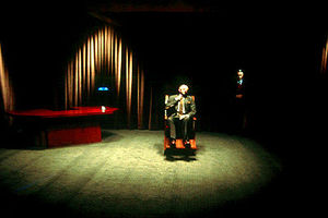 Michael J. Anderson seated in a large wooden wheelchair under an intense beam of light in a large and sparsely furnished room; a desk is in a far corner and the walls are covered in curtains