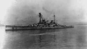 A large gray warship sits motionless in harbor. Several other ships are faintly visible in the distance.