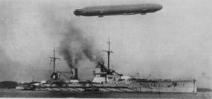 A large gray warship in harbor, a smaller boat is alongside. Thin smoke drifts slowly out of the two funnels.