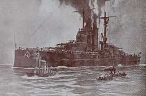 A large battleship sits motionless in the water with smoke coming out of it's funnels and three small boats moveing beside her in the foreground.