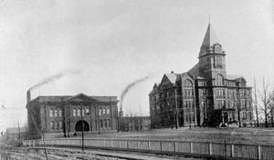 Two buildings stand side-by-side on a hill. The one on the left is two stories, with two smokestacks behind it. The one on the right is the larger, taller Tech Tower building.