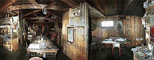  Dimly lit interior of a timber structure, with (left) a long table on which are papers and other unidentifiable objects. There are also bunks, sheles and packing cases. To the right, under a small window, is a smaller table with a chair.