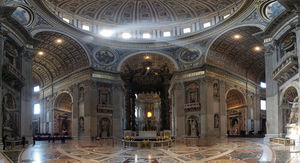 Wide angle photo of interior. Details visible in this view include: a golden band of text that extends around the cornice, niches with statues in every pier, carved angels around the arches, and the inlaid marble floor, the design of which radiates from the baldachin under the dome.