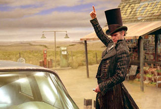 The character Count Olaf, played by Jim Carrey, sports an idyllic top hat and black-striped overcoat, seen from his thighs and up.  Behind him shows a bleak cornfield on the left and a gas station on the right.  The station has a red- and white-striped awning and looks worn.   Olaf faces to the driver's side window of a car, and he positions his right hand as if he's pointing at something above him, while concentrating his eyes on the window and holding car keys in his left hand.