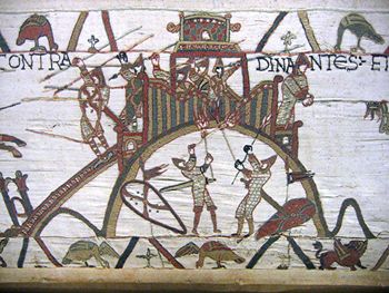 A section of an embroidered cloth showing a castle on a hilltop being defended by soldiers with spears while two soldiers in armour are attempting to set fire to the palisade