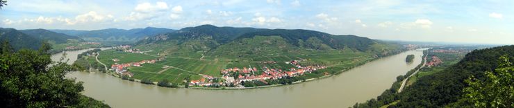 A vast river curves through a flat landscape. At the end of the flood plain, steep mountains rise above red-roofed houses.