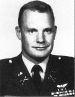 Head and shoulders of a white man with short hair and a slight smile, wearing a military jacket with ribbon bars and a winged pin on the left breast.