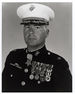 Head and shoulders of a middle-aged white man wearing a white peaked cap with oak leaf decorations on the visor and a dark military jacket with two rows of medals hanging from ribbons pinned to his chest and another medal hanging from his neck.