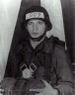 A black and white photo depicting Lozada from the waist up holding a sleepeing back in his hands, wearing a parachute and a Kevlar helmet with the letters A09 on it.