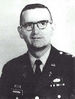 Head and shoulders of a middle-aged man with closely cropped hair and horn-rimmed glasses. He is wearing a dark military jacket with a parachute badge on the left breast and a Christian cross under a "U.S." pin on each lapel.