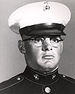 Head of a heavyset young white man with wire-frame glasses wearing a white peaked cap with black visor and a dark military jacket.