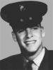 Head of a smiling young white man wearing a dark peaked cap and a dark jacket over a shirt and tie.