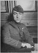 Head and torso of a man turned in his chair, his right arm laying stiffly on the arm rest in front of him. He is wearing a military uniform with a strap diagonally across the chest, a winged emblem on his left breast, and a garrison cap.