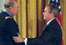 Profiles of two men shaking hands. The man on the right, wearing a dark suit, has his left hand on the other man's shoulder. The man on the left is taller and wearing glasses, a military jacket, and a light-blue ribbon around his neck.