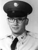 A black and white image of McWethy wearing military dress uniform with necktie and hat. He is wearing glasses and smiling at the camera.