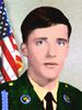 Head of a young white man with dark brown hair and blue eyes wearing a green military jacket. Behind him is an American flag.