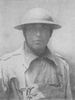 Head and shoulders of a man in a military coat, buttoned up to the neck, with a light pack strapped to his chest. He is wearing a metal helmet with a wide brim, its chin strap tightly cinched.