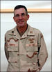 A color image of Roberts from the waist up in his desert camoflouge uniform. He is smiling with his hands crossed behind him and there is a dry erase board in the background.