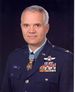 Head and shoulders of a white man with short gray hair, wearing a dark blue military jacket with rows of ribbon bars and a winged pin on the left breast, and a star–shaped medal hanging from a light blue ribbon around his neck.