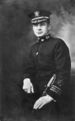 Head and torso of a man in a dressy military uniform, with white gloves, stripes on his sleeve near the wrist, emblems on his collar, a decorated peaked cap, and a sword held loosely in his hand.