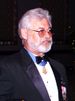 Head and shoulders of a man with gray hair, a full gray beard, and tinted glasses in a tuxedo. A medal, hanging from a blue ribbon around his neck, sits just below his bow tie.