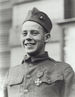 Head and shoulders of a happy young man in a military uniform with a single medal on his chest. He is smiling and his garrison cap is pushed high up on his forehead.