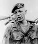 Head and shoulders of a white man wearing a t-shirt and a military jacket, unbuttoned at the collar, with the word "Rabel" on his right breast and "U.S. Army" on his left. He has on a beret and is holding a rifle over his shoulder by its barrel.