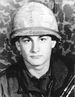 Head and shoulders of a young white man wearing a helmet with a camouflage-patterned cloth cover and a coat, standing before a camouflage-patterned background.