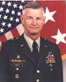 Head and torso of a white man with dark hair, gray on the sides, wearing a military jacket with pins a ribbon bars on the chest, lapels, and shoulders. A large American flag covers the background.