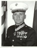 alt=A black and white photo of Modrzejewski from the waist up in his military dress blue uniform with hat and medals. There is an American flag and a Marine Corps flag in the background and his Medal of Honor is around his neck.