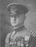 Head and shoulders of a white man in a military uniform with a long row of medals across his left breast, a strap laying diagonally across his chest, a high, stiff, collar, and a peaked cap.